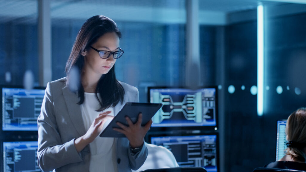 Young Female Government Employee Wearing Glasses Uses Tablet in System Control Center. In the Background Her Coworkers are at Their Workspaces with many Displays Showing Valuable Data. Information protection and cyber security. Modern safety digital background